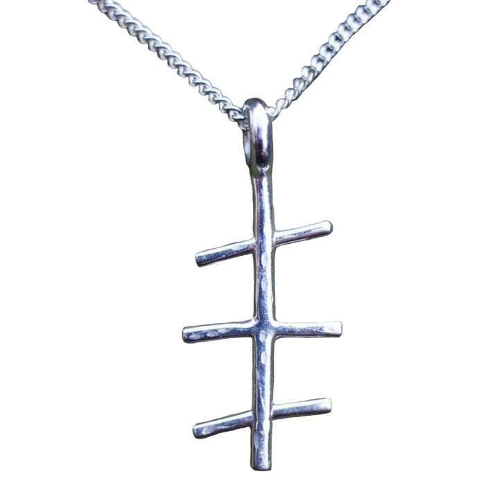 An Ogham Treasure Mam Small Pendant with a unique design resembling a vertical line intersected by three shorter horizontal lines hangs on a sterling silver chain. The background is moss-covered tree bark, creating a natural and rustic setting perfect for showcasing this exceptional Mothers Day gift.