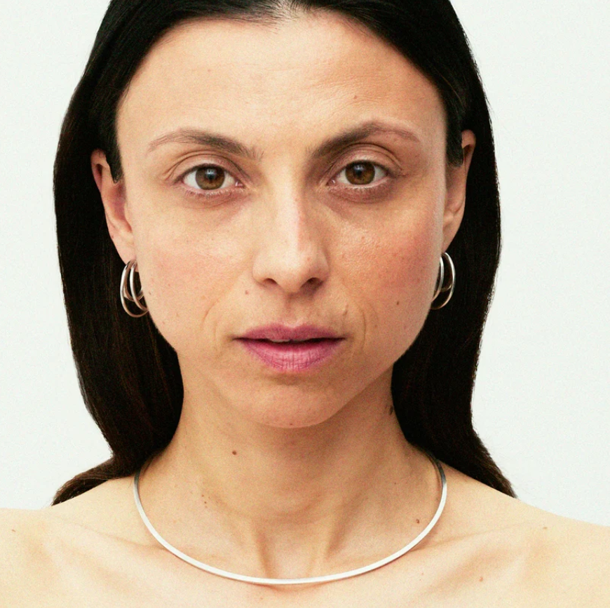 A close-up portrait of a woman with dark hair wearing silver hoop earrings against a plain light background.