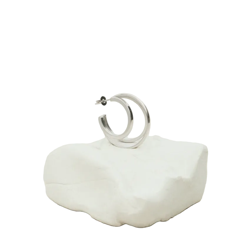 A polished silver hoop earring resting on a small white textured stone stand against a soft, light beige background.