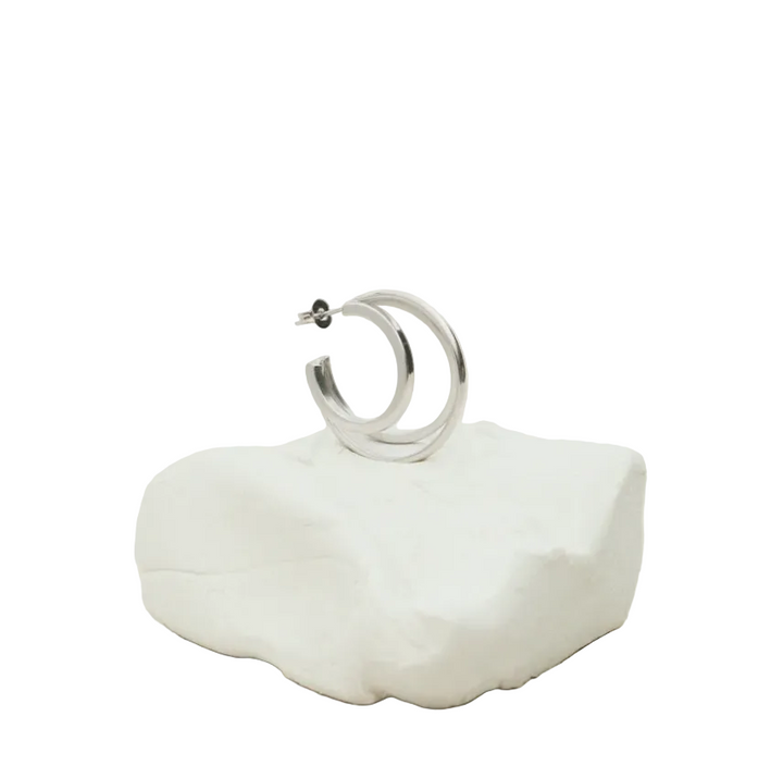 A polished silver hoop earring resting on a small white textured stone stand against a soft, light beige background.