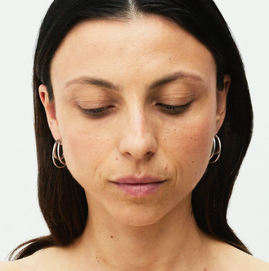Close-up of a woman with dark hair wearing simple gold hoop earrings against a plain light background.
