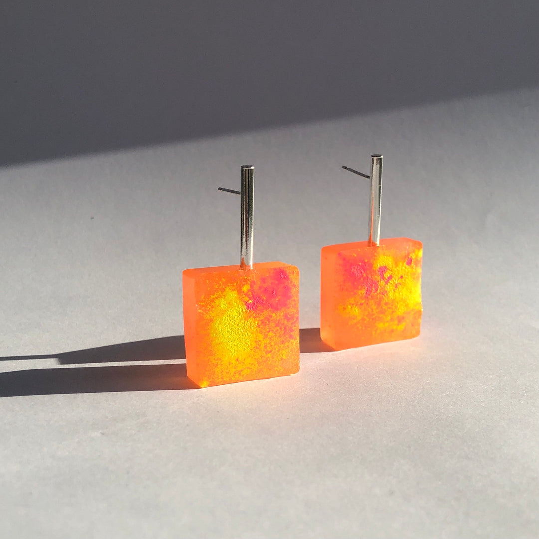 A pair of Angela O'Keefe Graffiti Block Earrings featuring square, neon orange and pink handmade resin blocks, each attached to a straight silver post. The earrings cast faint shadows on the light gray surface they are placed on, with sunlight highlighting their vibrant colors.