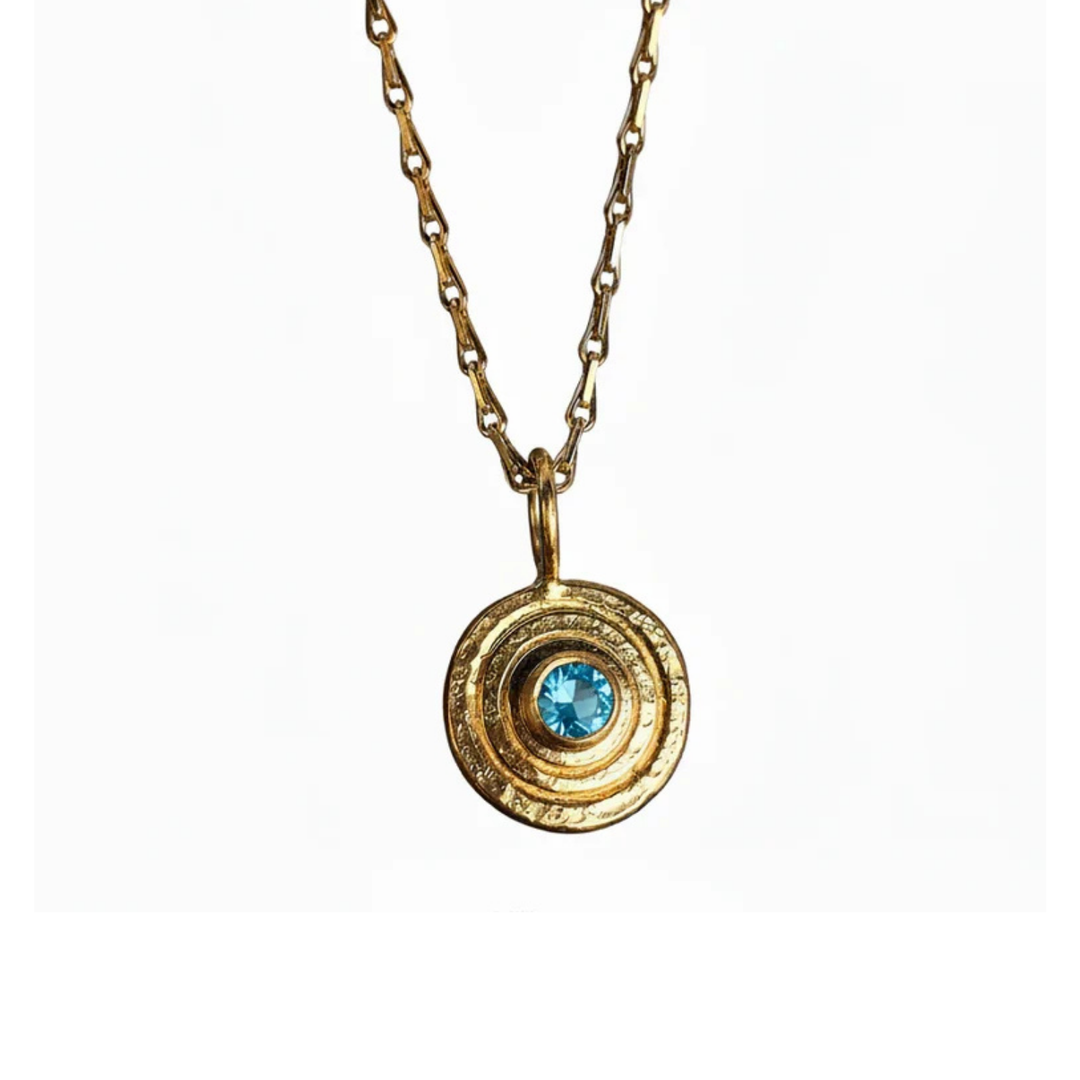 The "Orb Pendant Gold in Blue Topaz" by Caroline Stokesberry-lee features a gold-plated sterling silver chain with a circular pendant of concentric rings and a central blue topaz, shown against a white background.