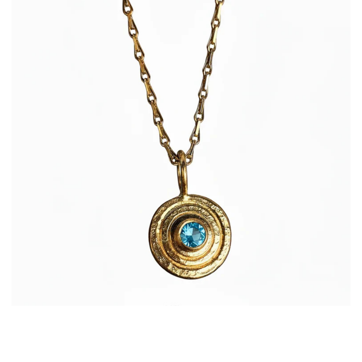 The "Orb Pendant Gold in Blue Topaz" by Caroline Stokesberry-lee features a gold-plated sterling silver chain with a circular pendant of concentric rings and a central blue topaz, shown against a white background.