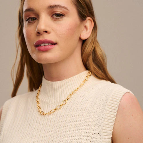 A person with long, light brown hair is wearing a sleeveless, cream-colored knitted top and an 18k gold plated Clean Oval Link Necklace - Gold by Orelia London. Their expression is neutral, and they are looking slightly to the side against a plain, light-colored background.