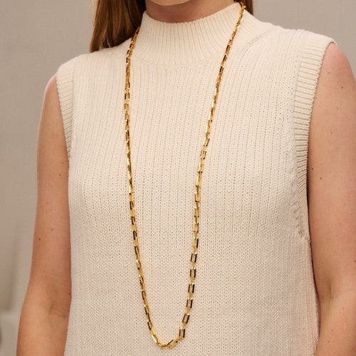 A person wearing a sleeveless, beige knitted top with a high neckline. Around their neck hangs Orelia London's Geo Rectangular Link Long Necklace With Clip Fastening - Gold. The background is neutral and out of focus.