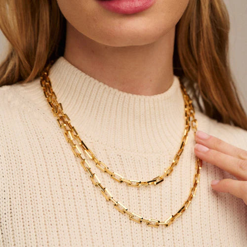 A person with light skin wearing a cream-colored ribbed sweater is shown from the chin down. They are wearing an Orelia London Geo Rectangular Link Long Necklace With Clip Fastening - Gold, and are touching the lower chain with their right hand, which has neatly manicured nails.