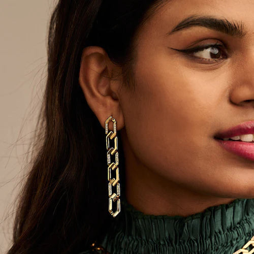 Close-up of a person wearing Orelia London Pave & Metal Chain Link Earrings - Gold with crystal pave encrusting. The person has winged eyeliner and pink lipstick. They are dressed in a green, high-neck garment, and the background is a neutral beige color.