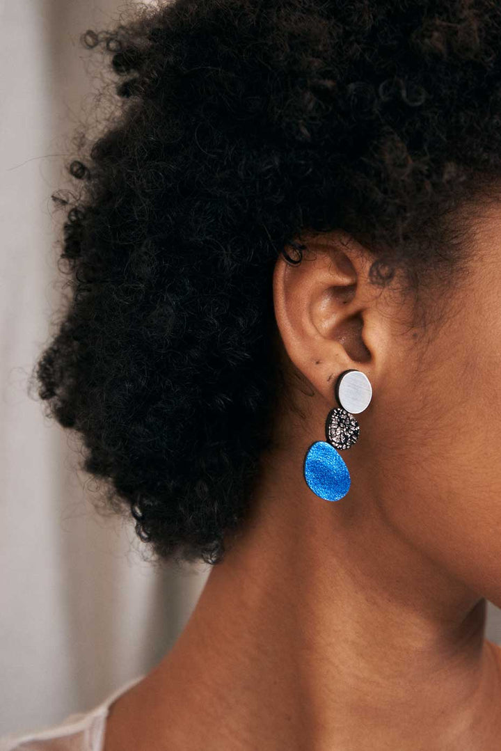 A close-up side view of a person with short, curly hair wearing Iskin Sisters' Organic Reflections Earrings. The earrings feature three elements: a small white circular section at the top, a black textured leather and acrylic middle section, and a larger bright blue circular section at the bottom.