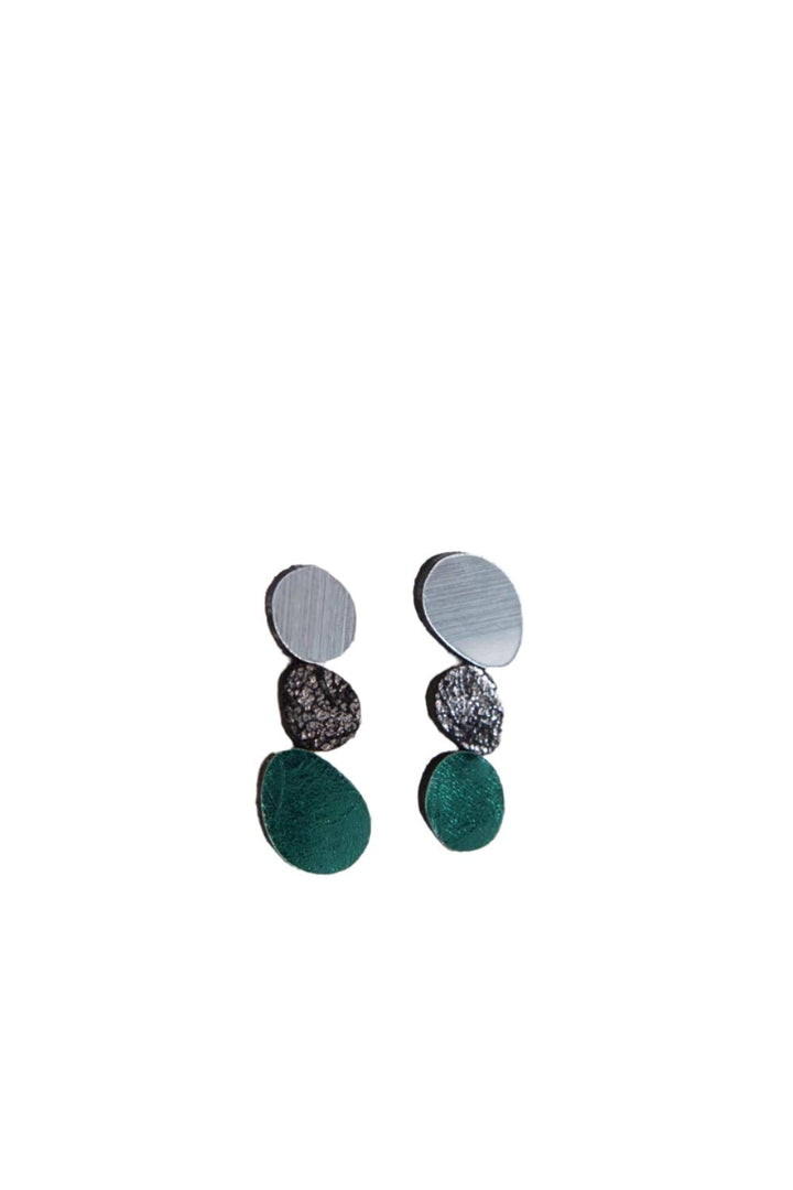 A pair of Organic Reflections Earrings by Iskin Sisters features three circular segments each: a silver top segment, a textured black leather middle segment, and a green acrylic bottom segment. The earrings are displayed against a plain white background.