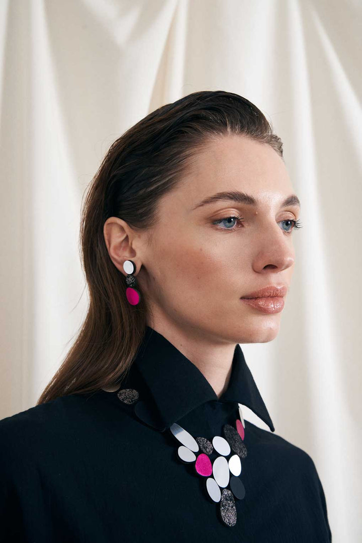 A close-up of a person with straight dark hair wearing a black collared shirt. They have on pink and silver geometric earrings from the Organic Reflections Earrings by Iskin Sisters, along with a matching necklace featuring circular and oval shapes. A white, slightly textured backdrop is behind them.