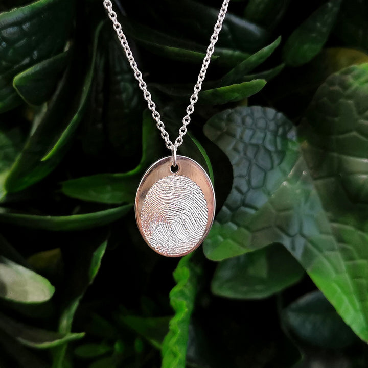 Displayed against lush green leaves, the Engraved Fingerprint Pendant by Personalised Charms is a silver necklace featuring a customized round pendant with a unique fingerprint design.