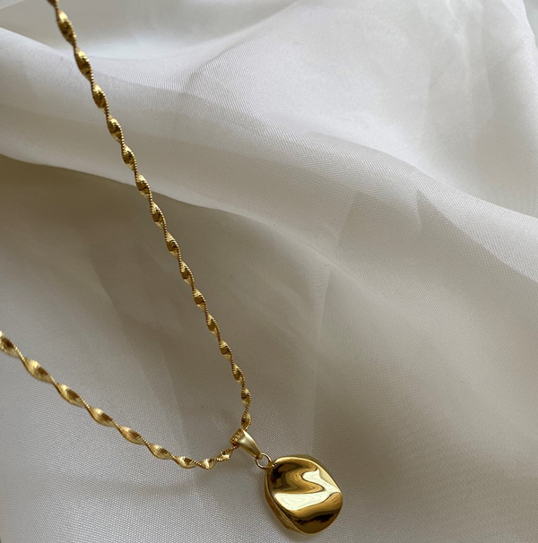 The Ovio Necklace by Bonvo, available in gold or silver, lies elegantly on a gently crumpled white fabric background. This piece features an 18ct gold plated twisted rope chain made of solid 925 sterling silver, adorned with a small hand-shaped pendant showcasing a smooth, irregular design. Soft lighting enhances the polished surface of the pendant and highlights the textural details of the chain.