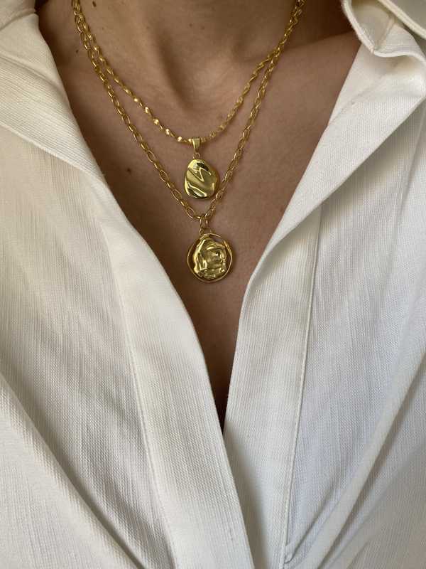 A person wearing a white, textured blouse showcases two Ovio Necklaces from Bonvo, layered with 18ct gold plating. One hand-shaped pendant displays an abstract design, while the other features a profile portrait engraving. The blouse’s collar and front are visible, emphasizing the elegance of the jewelry.