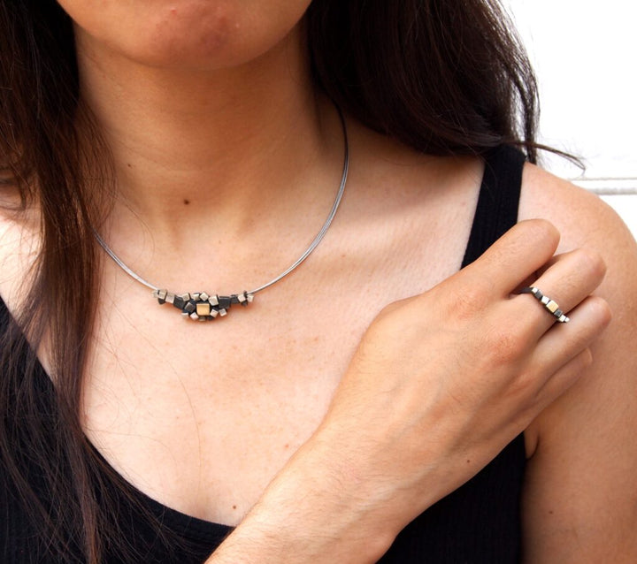 Close-up of a person wearing a black sleeveless top. They have long dark hair and are wearing a metallic necklace with geometric bead details from the Iceland collection. Their right hand, adorned with a matching Matt Silver Narrow Iceland Ring from MarCucurella, rests near their collarbone. The background is neutral.