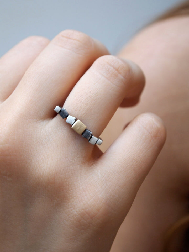 Close-up of a hand wearing a MarCucurella Narrow Iceland Ring with alternating metallic cube-shaped segments in matt silver and 18kt yellow gold tones. The ring is minimalistic with a modern design, part of the Iceland collection, highlighting the unique pattern of the cubes. The background is blurred, focusing on the ring detail.