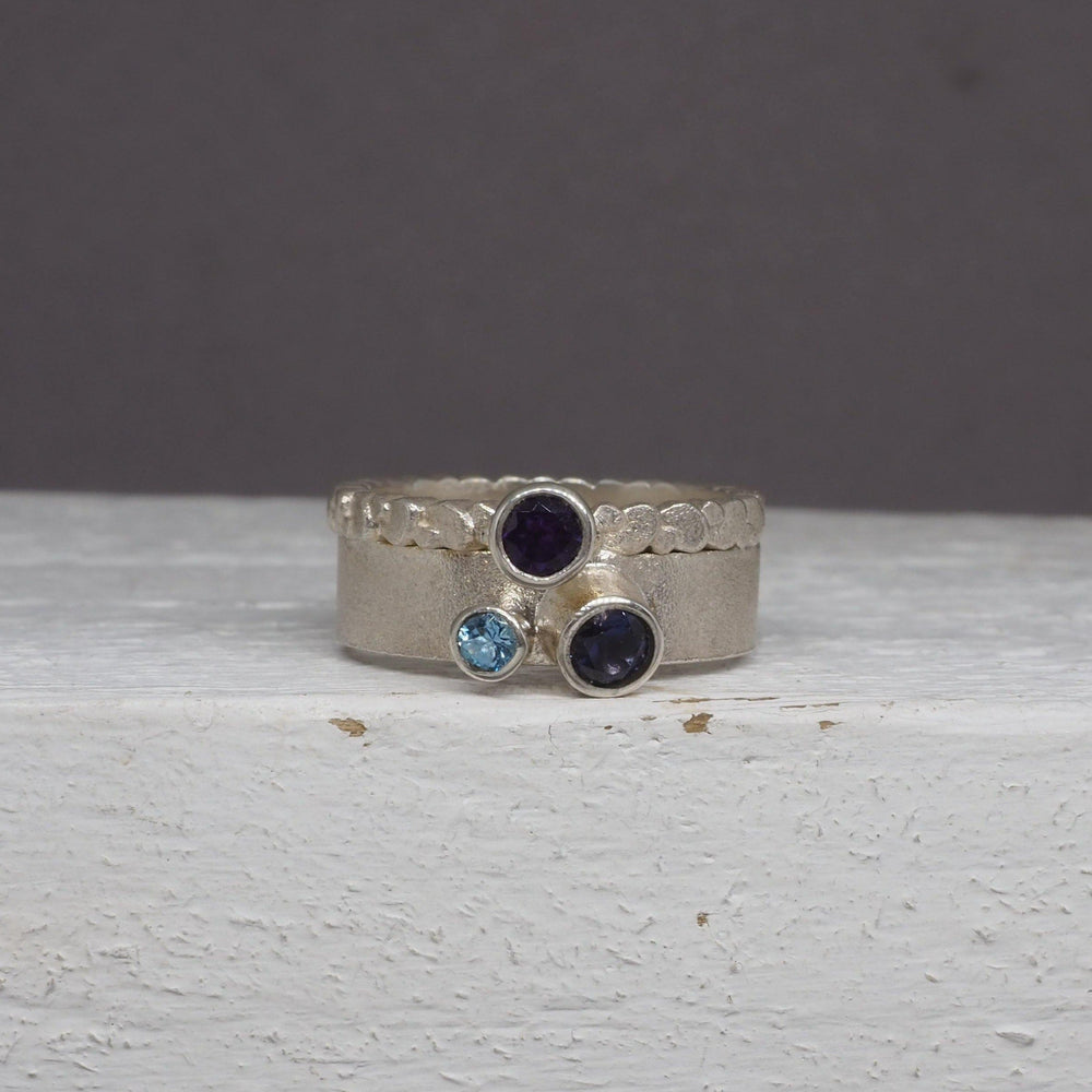 A frosted silver ring with an etched design is set with one small blue gemstone and two larger purple faceted stones. The Iolite, Amethyst & Aqua Marine Wing Ring by Leoma Drew is displayed on a light-colored textured surface against a plain gray background.