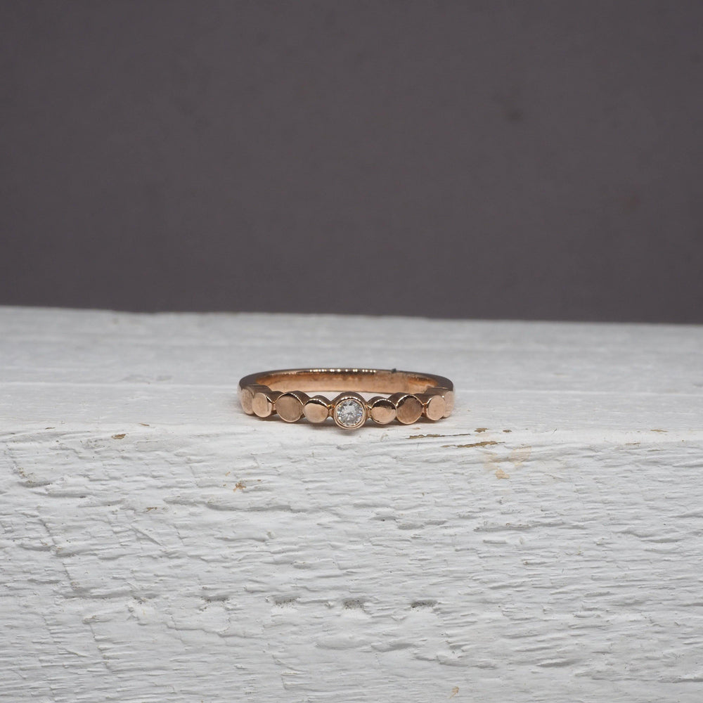 A delicate Flow Pebble Ring 9ct Rose Gold with Diamond by Miriam Wade rests on a white, wooden surface against a plain gray background. This exquisite piece is made to order.