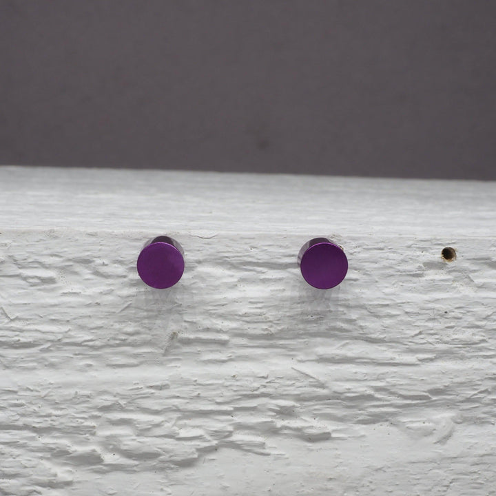 Two Cylinder Studs in a small purple hue, reminiscent of delicate stud earrings, are pinned to a white, textured surface with visible brush strokes. The surface has a slight overhang, and the background is muted grey. A small hole is visible to the right of the studs. These studs are part of the Filip Vanas collection.