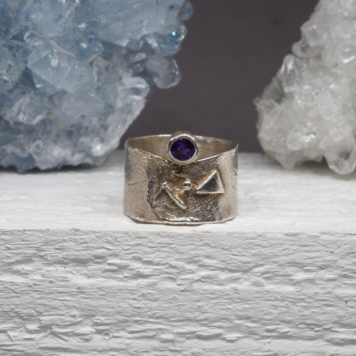 A sterling silver Amethyst Guardian Ring by Caroline Stokesberry-lee with a wide band sits on a white surface, decorated with engraved geometric shapes. The ring features a small, round Amethyst gemstone set in a bezel. In the background, there are clusters of light blue and white crystals.