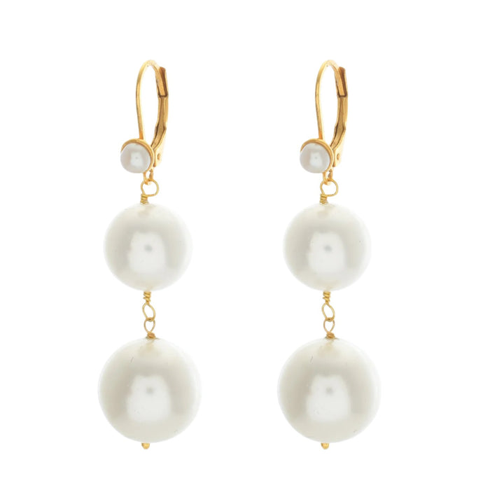 The Shyla Pernille Earrings feature two large white pearls stacked vertically, each topped with a small pearl and attached to a 22 carat gold lever-back hook. Separated by gold wire, these elegant earrings reflect a classic and sophisticated design.