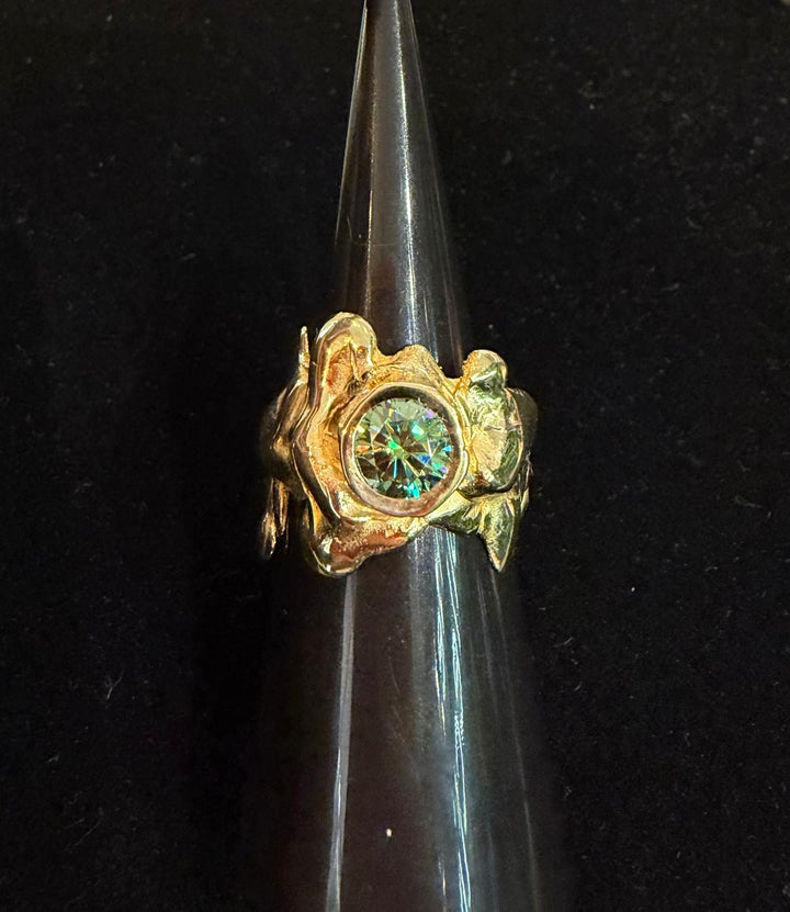 The Solstice Ring with Large Moissanite by Madeleine Sculptural Jewellery features a handcrafted, sculpted gold band and a round moissanite center, shown on a dark ring holder against a black background.