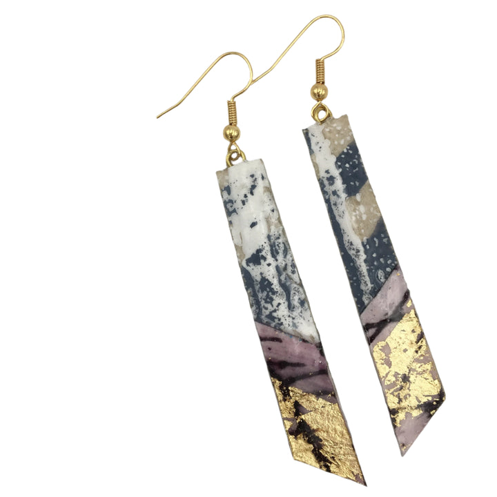 A pair of Panthea Batik Textile Earrings in Dove/Gold/Aubergine from Rothlú, featuring a marbled and abstract design with hypo-allergenic gold plated hooks, lies on a white surface next to some brown dried grass stems.