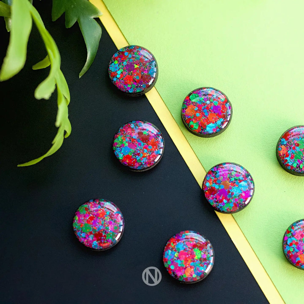 Eight colorful Party Stud Earrings by Naoi with red, blue, and green splatters are placed on a surface divided diagonally into lime green and black sections. Hand-painted wooden studs add a unique touch while green plant leaves in the upper left corner and a small logo at the bottom center complete the scene.