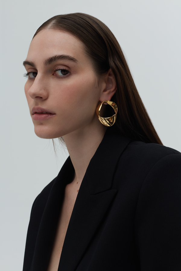 A woman with long, straight brown hair is wearing a black blazer and large, round gold-plated earrings. She has a neutral expression and is looking slightly to her left. The background is plain and light-colored.

Revised Sentence: A woman with long, straight brown hair is wearing a black blazer and Bonvo's Paula Curved Earrings (Small). She has a neutral expression and is looking slightly to her left. The background is plain and light-colored.