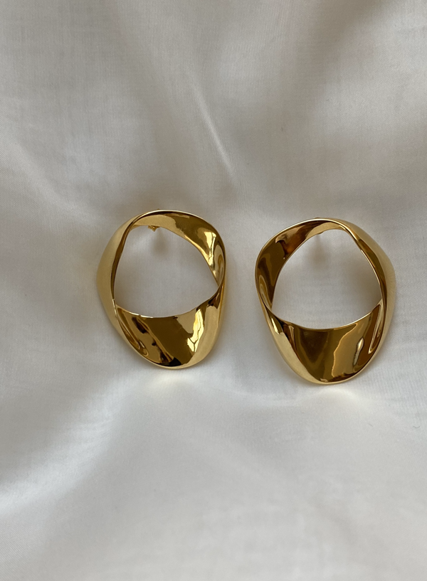 A pair of shiny gold plated hoop earrings with a twist design, resting on a soft white fabric background. The Paula Curved Earrings (Small) from Bonvo have a smooth, reflective surface that catches the light.