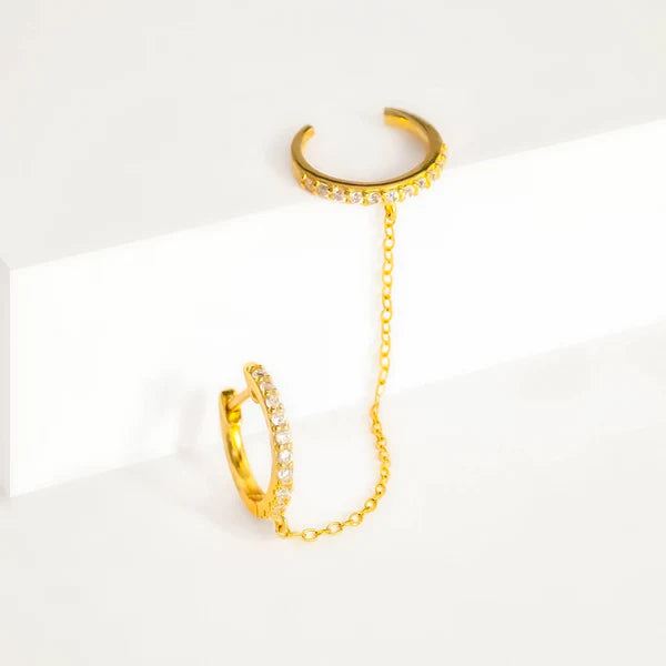 A pair of Pavé Huggie Hoop & Cuff by Marie June Jewellery connected by a delicate chain. Each cuff is adorned with small, sparkling clear gemstones, and the chain dangles elegantly between them. The handmade jewelry is displayed on a white surface, highlighting its simplicity and elegance.