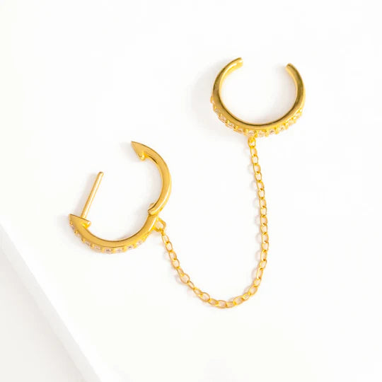 A close-up photo of a handmade, gold Pavé Huggie Hoop & Cuff by Marie June Jewellery. The earring consists of two small, open-ended hoops connected by a delicate 18K yellow gold chain. One hoop has a post for piercing, and both hoops are adorned with small, clear gemstones. It rests on a white surface.