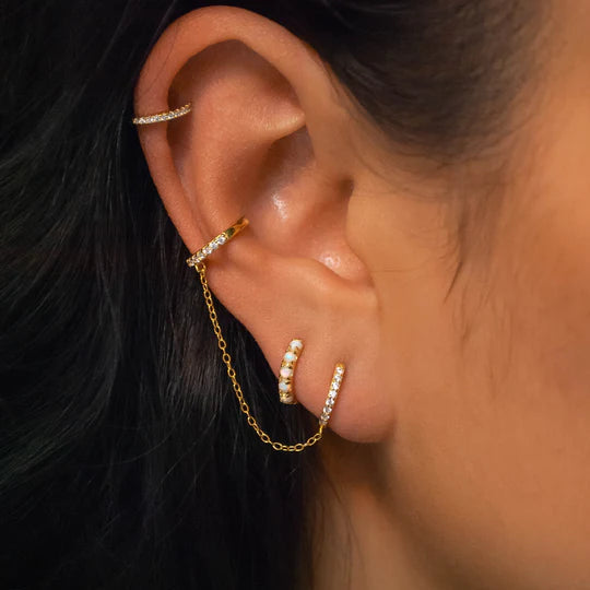 Close-up image of an ear adorned with multiple handmade gold earrings. There are three 18K yellow gold Pavé Huggie Hoop & Cuff by Marie June Jewellery connected by a delicate chain, all embellished with small white stones. Additionally, there are two curved bar piercings above the hoops adding to the ornate look.