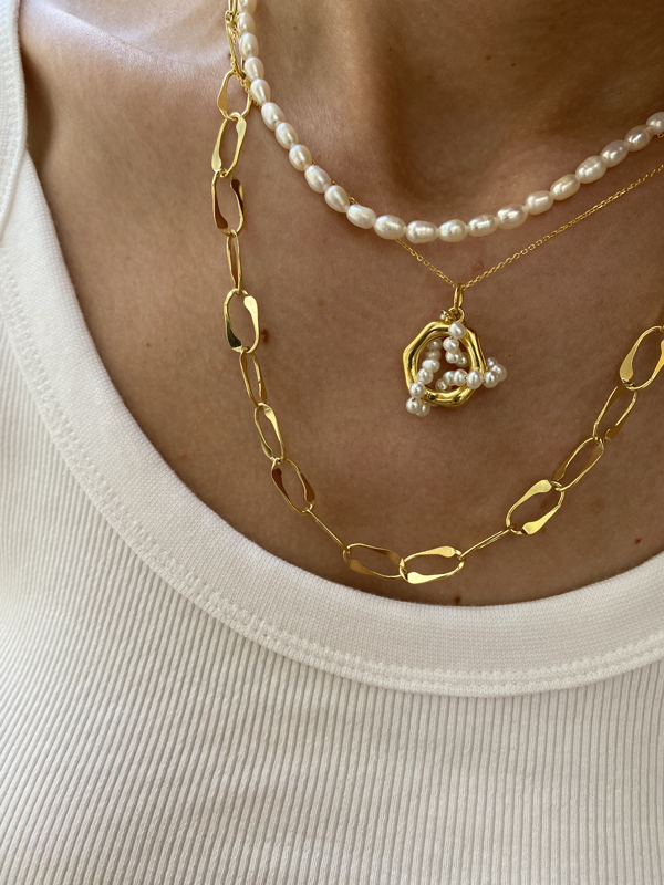 Close-up of a person wearing two necklaces over a white ribbed top. One necklace is a string of small freshwater pearls, and the other is the Bonvo Pearl Parade Necklace, an 18ct gold-plated chain with irregular, open links, featuring a cluster of pearls in a gold setting as a pendant.