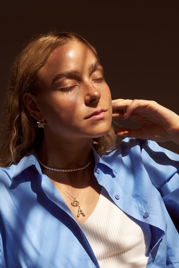 A woman with closed eyes basks in sunlight, casting shadows on her face. She has light brown wavy hair and wears a blue button-up shirt over a white top. She accessorizes with Bonvo's Pearl Parade Necklace, an "A" pendant in 925 sterling silver, and small earrings.