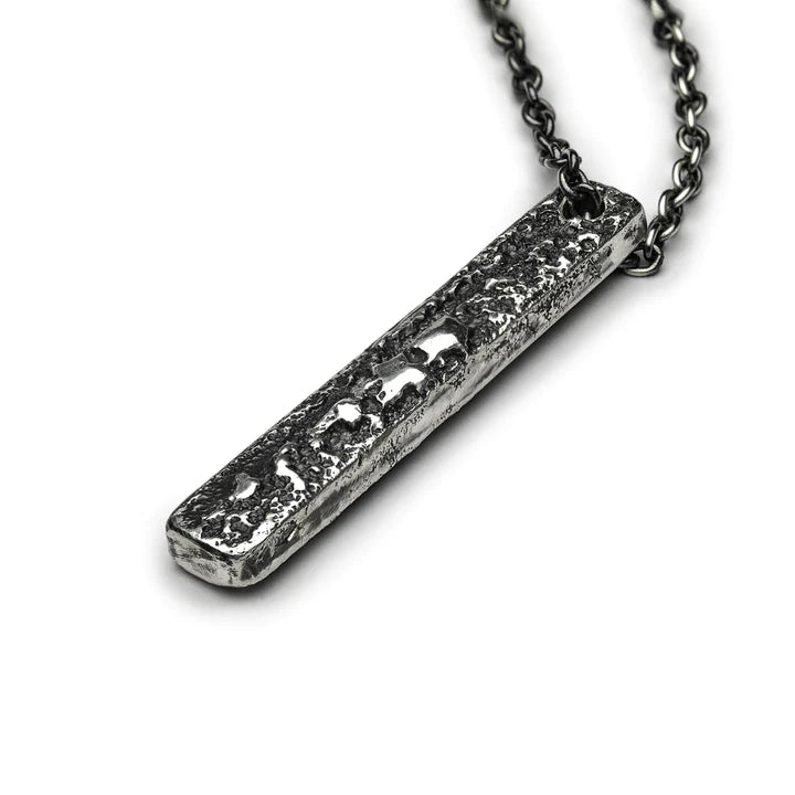 A rectangular, textured metal pendant hangs from a dark, robust chain. The patinated 925 silver pendant has a distressed, rugged surface, giving it an aged and rustic appearance. The chain is made up of interlocking links, complementing the pendant's rough aesthetic. This is the Slim Necklace (Regular) by Septentrion.