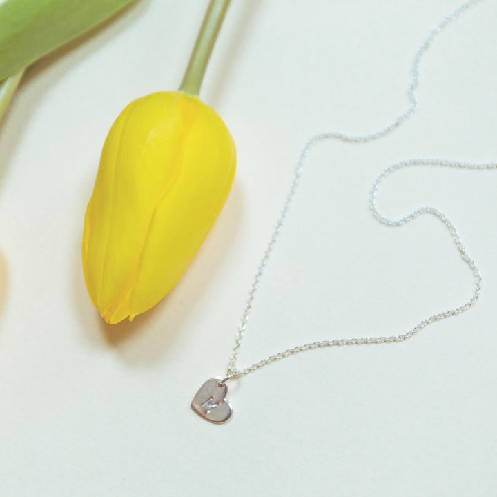 A delicate gold necklace with a small heart-shaped pendant lies on a white surface beside a vibrant yellow tulip. The Personalised Charms necklace, featuring an adjustable oval belcher chain, is spread out in a gentle curve, complementing the simplicity and elegance of the scene.