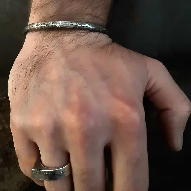 Close-up of an adult's right hand resting on a surface, adorned with handmade jewellery by Septentrion, including a patinated 925 silver bracelet and a Cursor Ring on the ring finger. The hand has a visible amount of hair, and the background is dark and blurred.