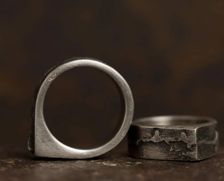 A close-up of two rustic, handmade Cursor Rings by Septentrion. The ring on the left has a smooth, thin band and stands upright, while the ring on the right, crafted from patinated 925 silver, has a thicker, textured band with a rectangular shape and lies flat on a dark surface.