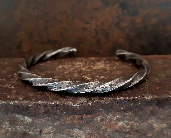 A rustic, twisted metal bangle bracelet rests on a weathered, brown and red, textured surface with a matching backdrop. The patinated 925 silver Twisted Bracelet by Septentrion features a simple yet rugged design with a dark, worn finish, giving it an artisanal and vintage appearance.