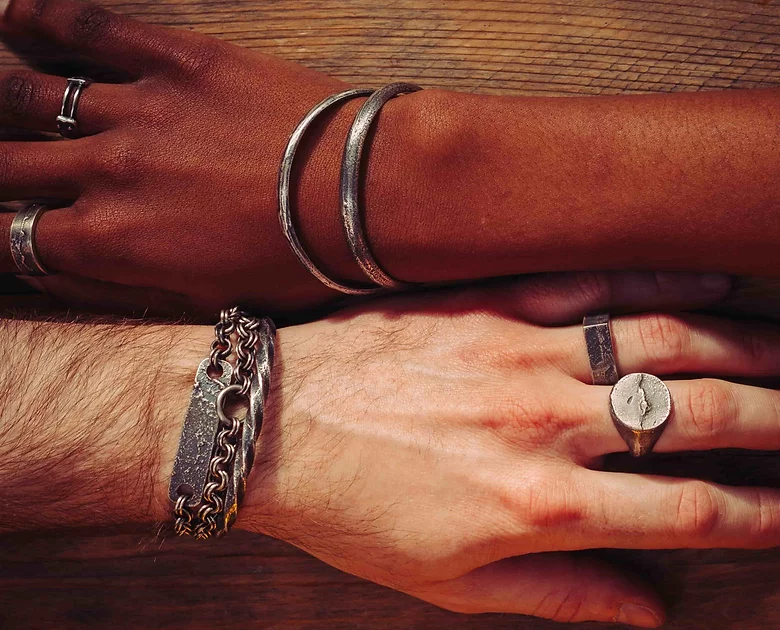 Two hands with various silver and metal accessories rest on a wooden surface. The upper hand, darker-skinned, wears two bangles of patinated 925 silver, while the lower hand, lighter-skinned, is adorned with handmade jewelry showcasing unique designs—a Septentrion Twisted Bracelet and several rings.