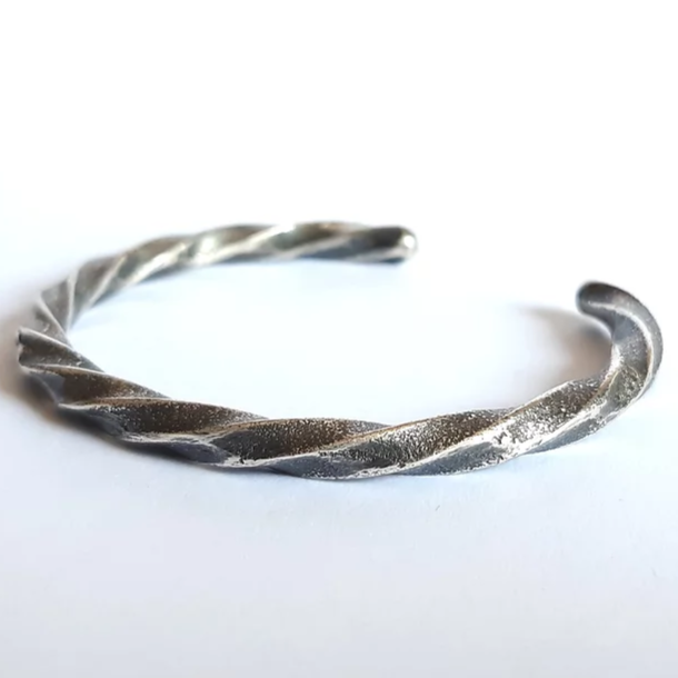 A twisted, textured patinated 925 silver **Septentrion Twisted Bracelet** with an open-ended design, resting on a white surface. The bracelet features a helical pattern that gives it a modern yet rustic appearance, exemplifying the charm of handmade jewelry.