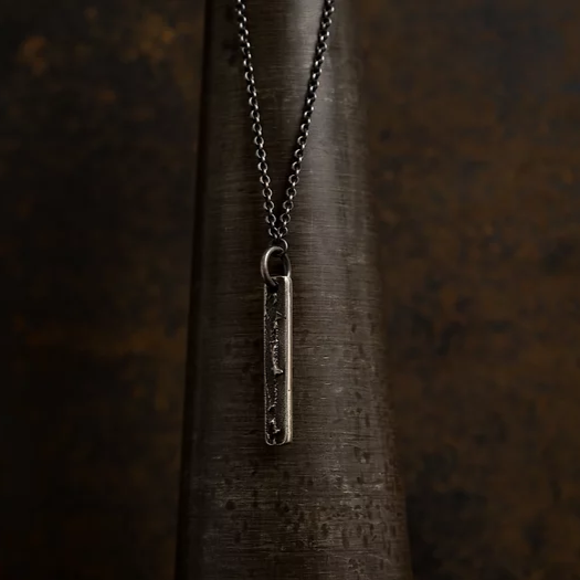 A close-up of a chain with a vertical, rectangular pendant in patinated 925 silver hangs against a dark, textured background. The handmade jewelry features unique patterns engraved along its length, and the chain is a simple, linked style. The lighting creates a somber, moody atmosphere. This is the Slim Necklace (Regular) by Septentrion.