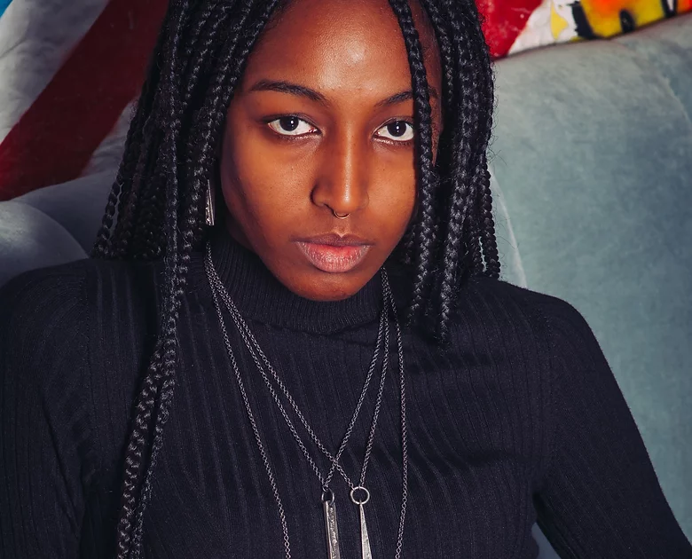 A person with long braided hair, wearing a black turtleneck sweater and multiple pieces of Septentrion's Slim Necklace (Regular), sits against a light-colored backdrop with unique patterns. The expression is neutral, with a focused gaze towards the camera.