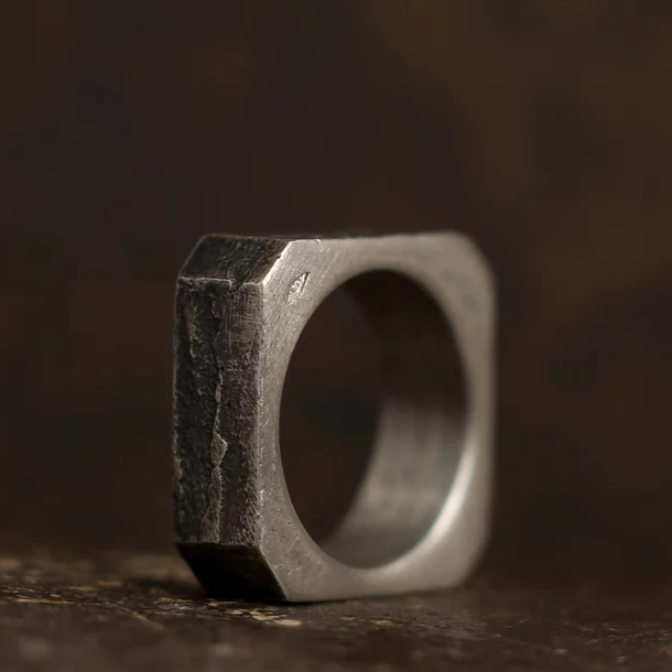 A close-up image of a rugged, square-shaped metal ring with a textured finish. Made from patinated 925 silver, the ring has visible signs of wear, giving it a rustic and industrial appearance. The background is out of focus, emphasizing Septentrion's Nut Ring unique form and surface details.