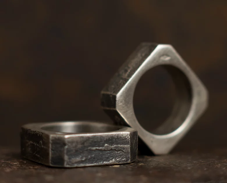 Close-up of two Septentrion Nut Rings with hexagonal shapes placed on a dark surface, resembling handmade jewellery. One Nut Ring is standing upright, while the other lies flat. The background is blurred and dark, emphasizing the unique patterns and texture of the rings.