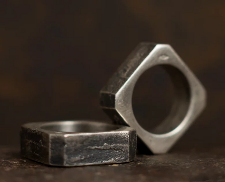 Close-up of two Septentrion Nut Rings with hexagonal shapes placed on a dark surface, resembling handmade jewellery. One Nut Ring is standing upright, while the other lies flat. The background is blurred and dark, emphasizing the unique patterns and texture of the rings.