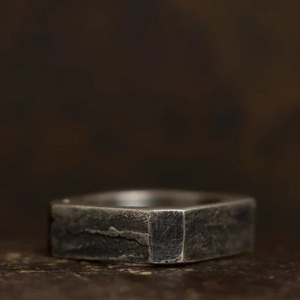 A close-up image of a square-shaped metal ring with a dark, rustic finish. Made from patinated 925 silver, the surface of the Nut Ring by Septentrion appears to have a worn and textured look, adding to its rugged aesthetic. The background is blurred and dark, making this piece of handmade jewellery stand out prominently.