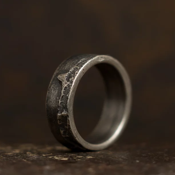 A close-up shot of a rustic, weathered metal ring set against a dark, blurred background. The Septentrion Wide Ribbon Ring has a rough, textured surface with unique patterns and uneven edges, giving it an antique and artisanal appearance.