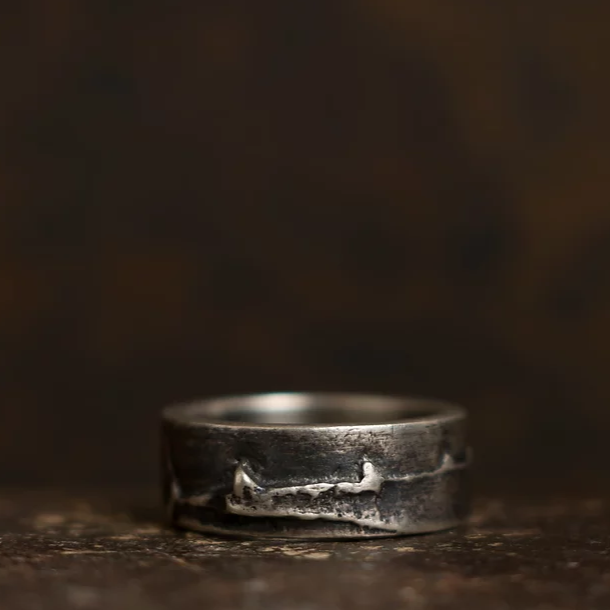 A close-up of a rugged metal ring with a worn texture and uneven surface, set against a dark, blurred background. The Wide Ribbon Ring from Septentrion has a handmade, rustic appearance with slight imperfections and unique patterns, giving it an artisanal quality.